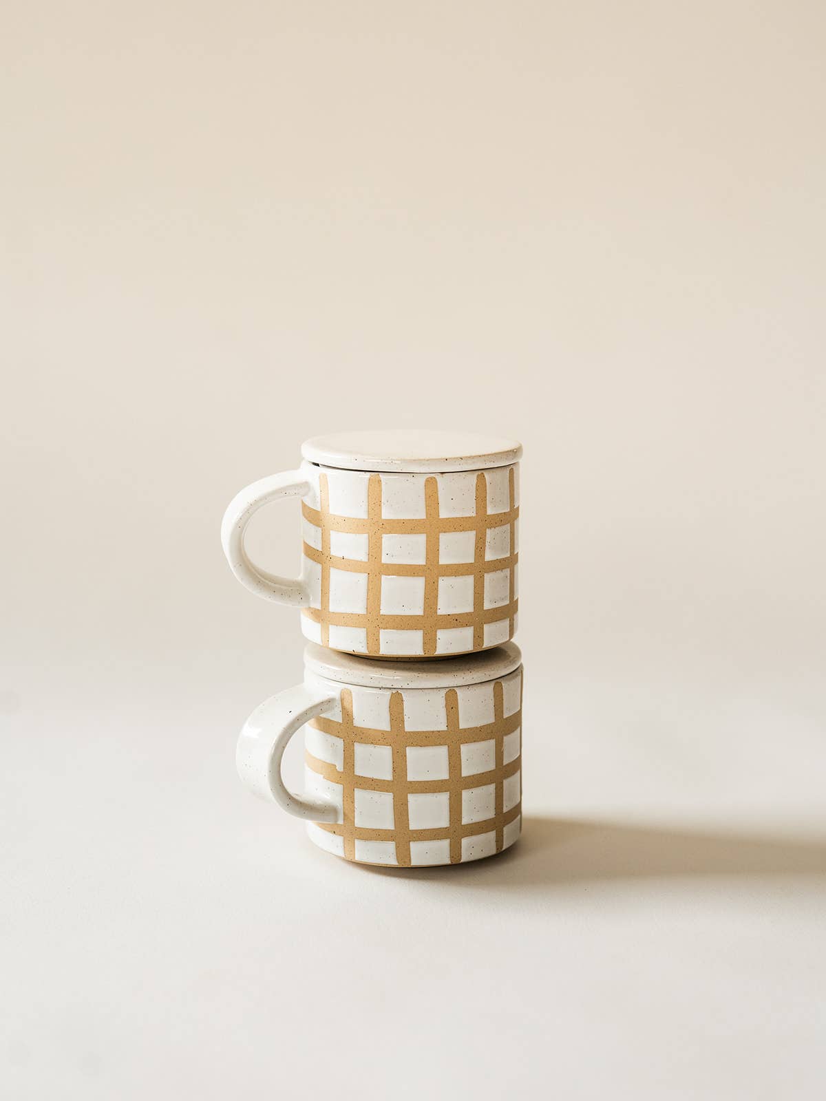 Two ceramic mugs with beige checkered pattern stacked on a light background